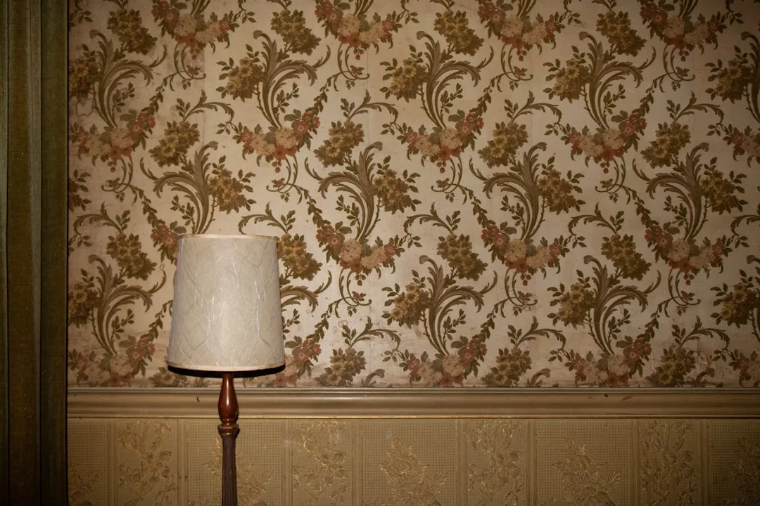Vintage floral wallpaper with ornate scrolling patterns on a wainscoted wall beside a classic floor lamp