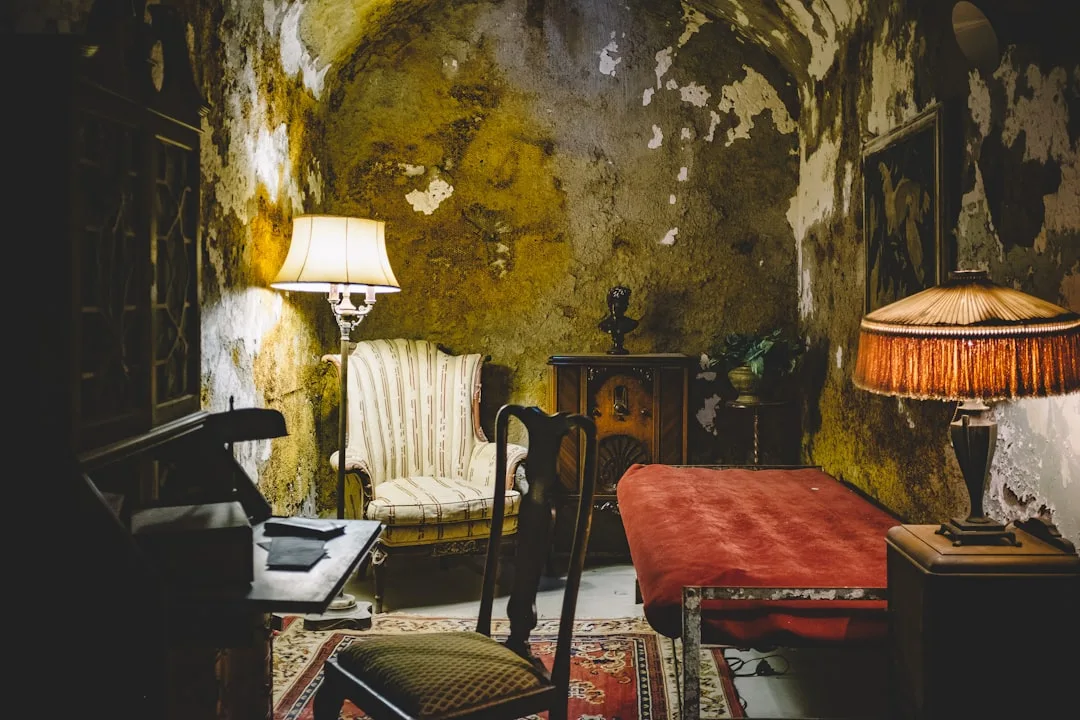 Abandoned grunge room with peeling walls, vintage furniture, antique lamps and worn red velvet table in dim moody lighti