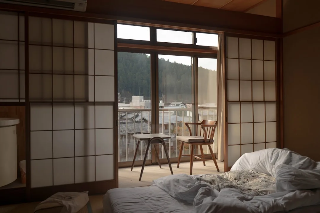 Japanese-style bedroom with shoji sliding doors, wooden chairs, and unmade bed overlooking rooftops and forested hills