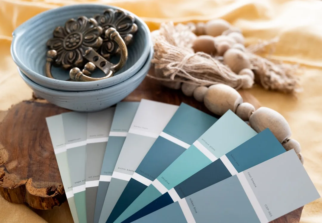 Paint color swatches in blue-gray and warm neutral tones fanned out on rustic wood surface with decorative bowl and bead