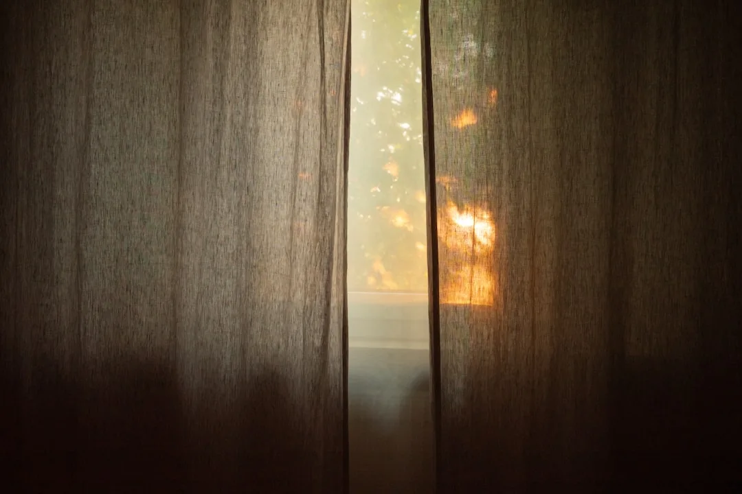 Sheer linen curtains partially open revealing warm sunset light filtering through window with outdoor trees visible