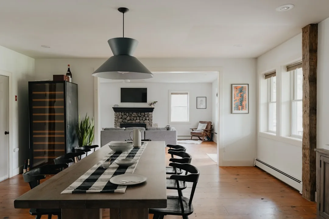 Modern farmhouse dining room with wood table, black pendant light, plaid runner, and stone fireplace living room beyond