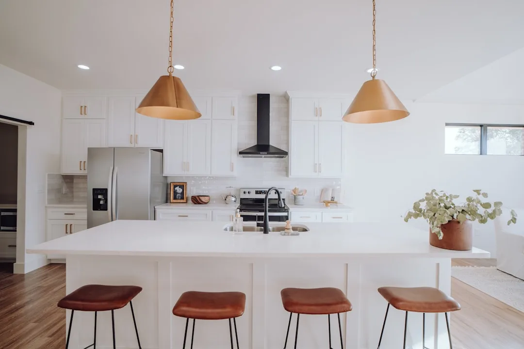 Modern white kitchen with gold pendant lights, white quartz island, brown leather bar stools, and stainless steel applia