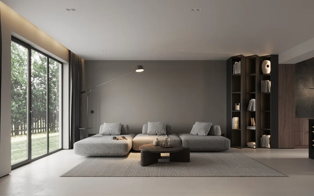 Minimalist living room with gray sectional sofa, dark wood bookshelf, concrete floors, and floor-to-ceiling windows