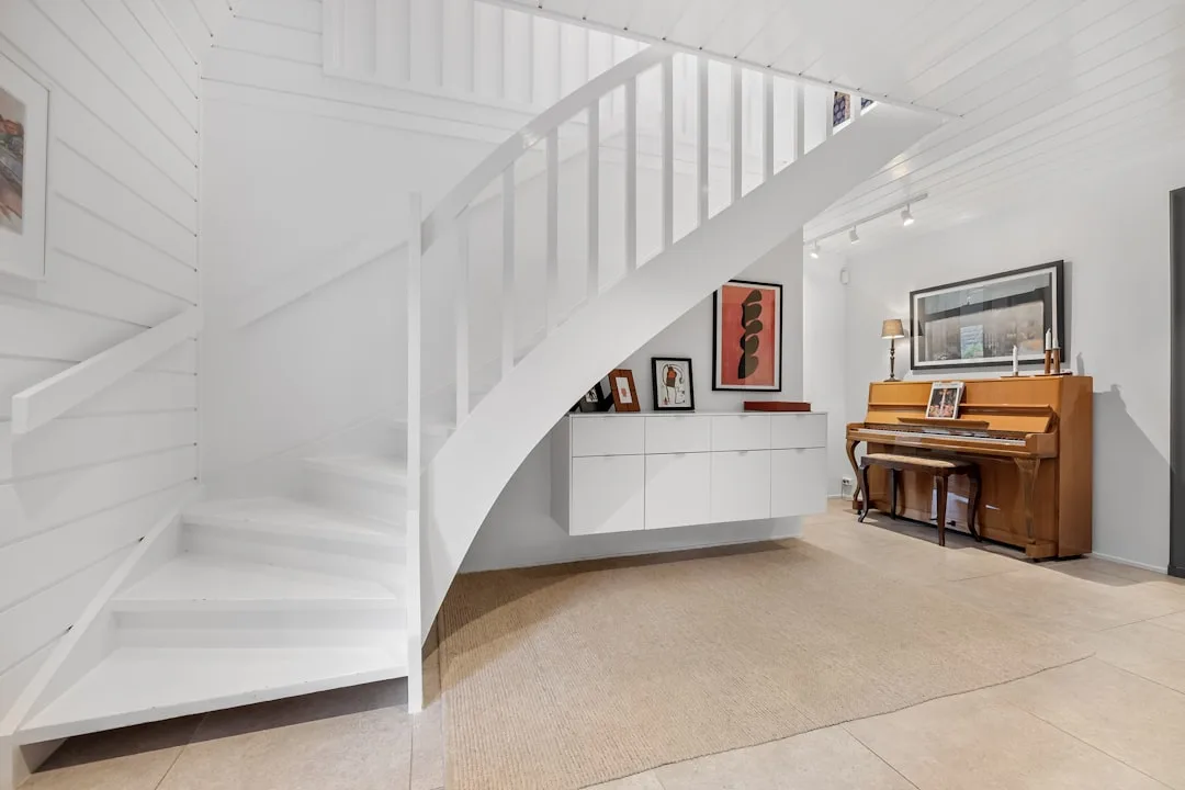 White floating cabinet built under staircase with artwork display next to wooden upright piano in modern home