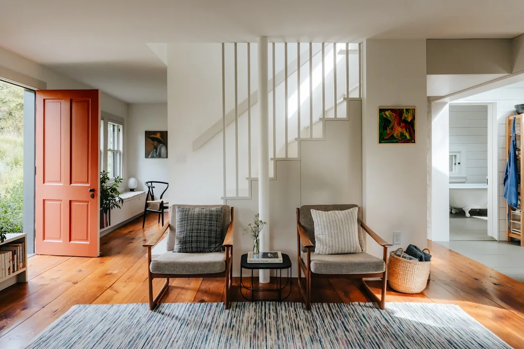 Bright mid-century modern living room with wood floors, two gray armchairs, white staircase, and orange front door