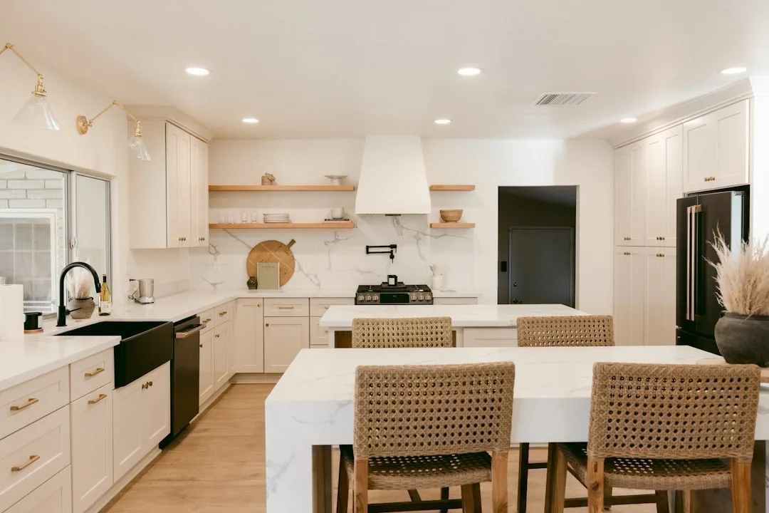 High-end white kitchen remodel with marble island, open shelving, black appliances, and rattan bar stools