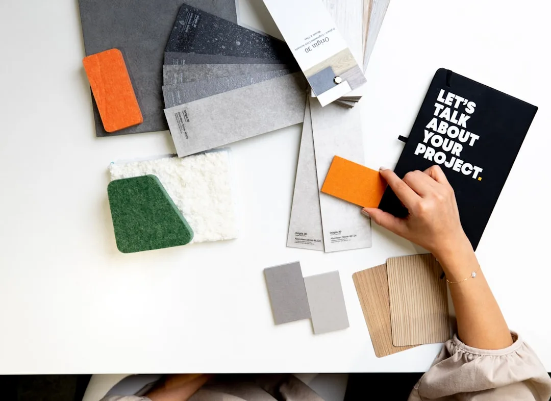 Hand holding material samples and black notebook saying Let's Talk About Your Project on white desk with wallpaper swatc