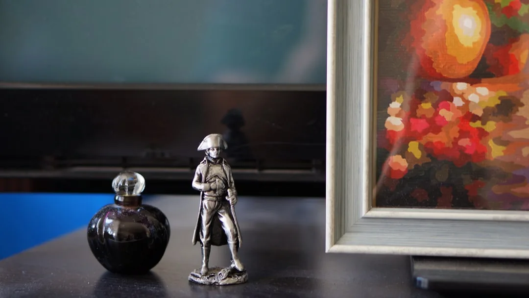 Small metal Napoleon figurine beside a dark glass perfume bottle and framed painting on a shelf