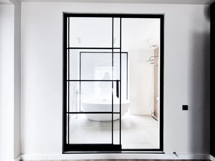 Black steel-framed glass pocket door sliding open to reveal a modern white freestanding bathtub bathroom