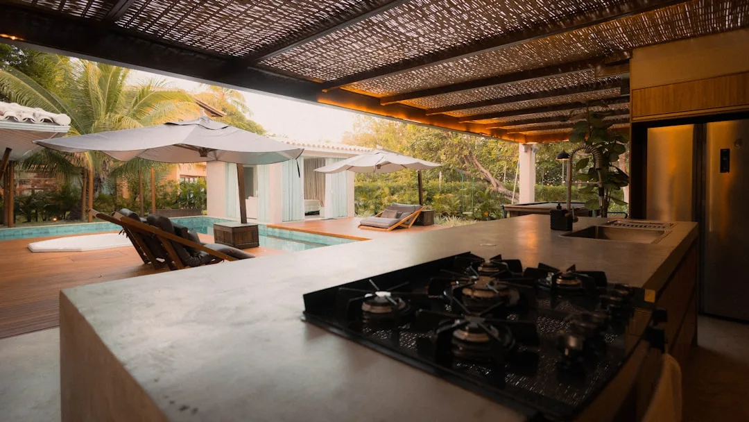 Modern outdoor kitchen with gas cooktop and concrete island overlooking a luxury pool and pergola in tropical setting