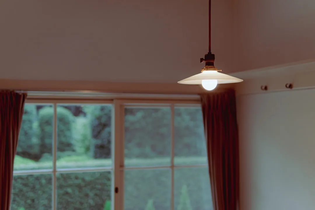 Pendant light with white shade hanging from ceiling in small room with large window and dark curtains