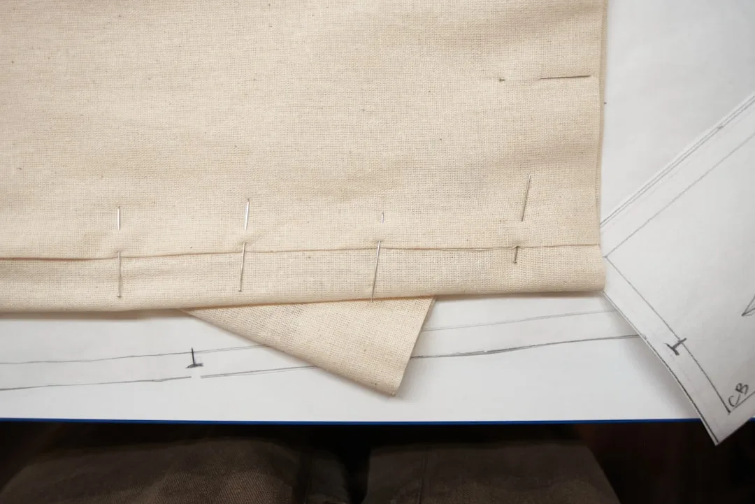 Curtain panel side hem pinned with sewing pins on fabric laid over paper pattern showing hem measurement lines