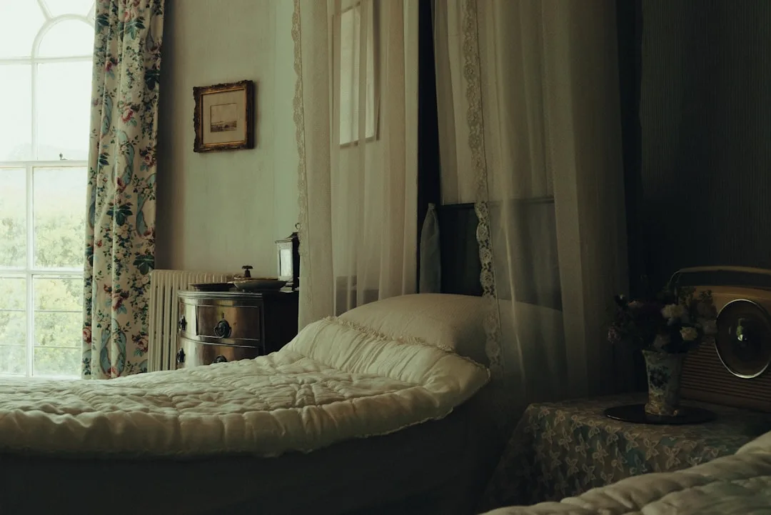 Vintage guest bedroom with layered white quilted bedding, lace canopy curtains, floral drapes, and antique nightstand