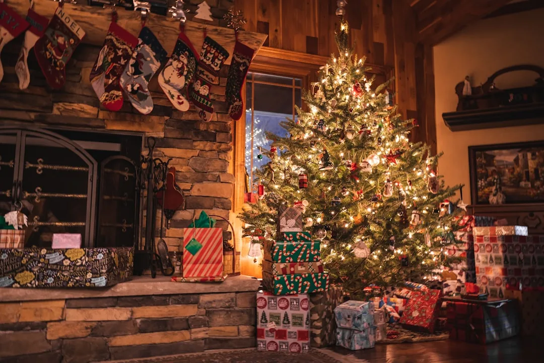 Decorated Christmas tree with lights and wrapped presents next to stone fireplace with hanging stockings in cozy living 