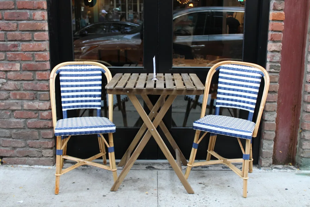 Freestanding bistro setup with two blue-and-white wicker chairs and folding wooden table on outdoor cafe sidewalk