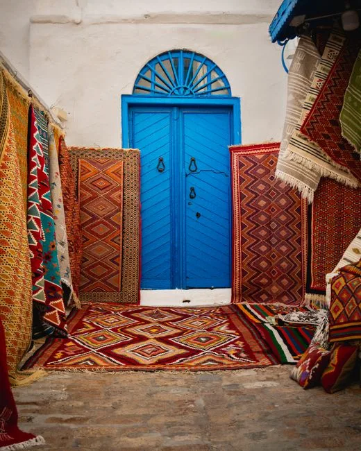 Colorful layered Moroccan rugs displayed in front of a vibrant blue arched door in a traditional market