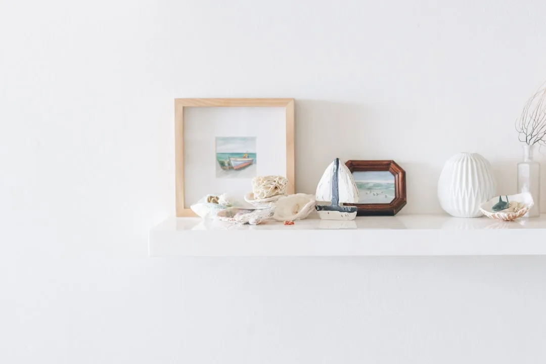 White floating shelf styled with framed art, wooden sailboat, seashells and white ceramic vase in minimalist coastal dec