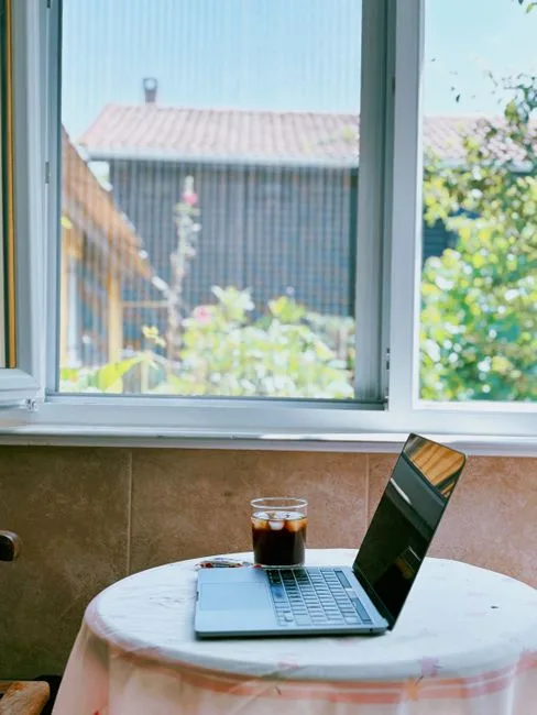 Laptop and iced coffee on small table by window overlooking lush garden, indoor workspace with outdoor view