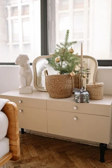 Small potted Christmas tree in wicker basket on beige dresser with candle and holiday gift in cozy apartment