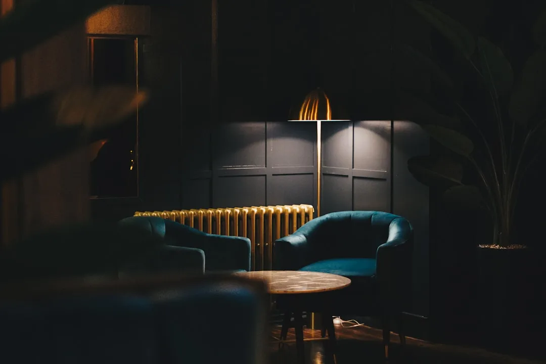 Gold floor lamp casting warm light in dark lounge with teal velvet chairs, demonstrating statement lighting as a zone si