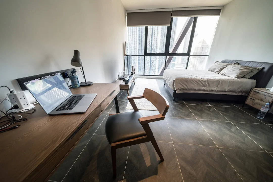 Modern bedroom with built-in wooden desk, open laptop, and chair integrated alongside a bed with city views