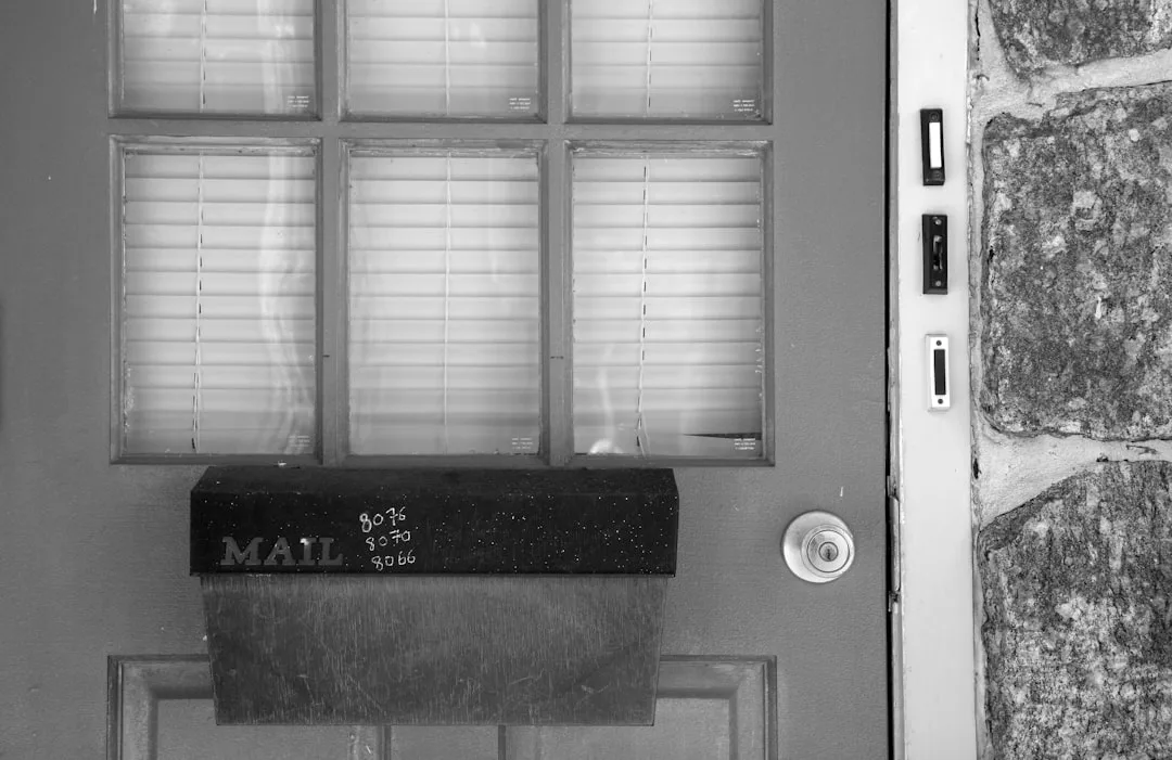 Black and white photo of a front door entryway with mail slot, window blinds, doorbell, and stone wall detail