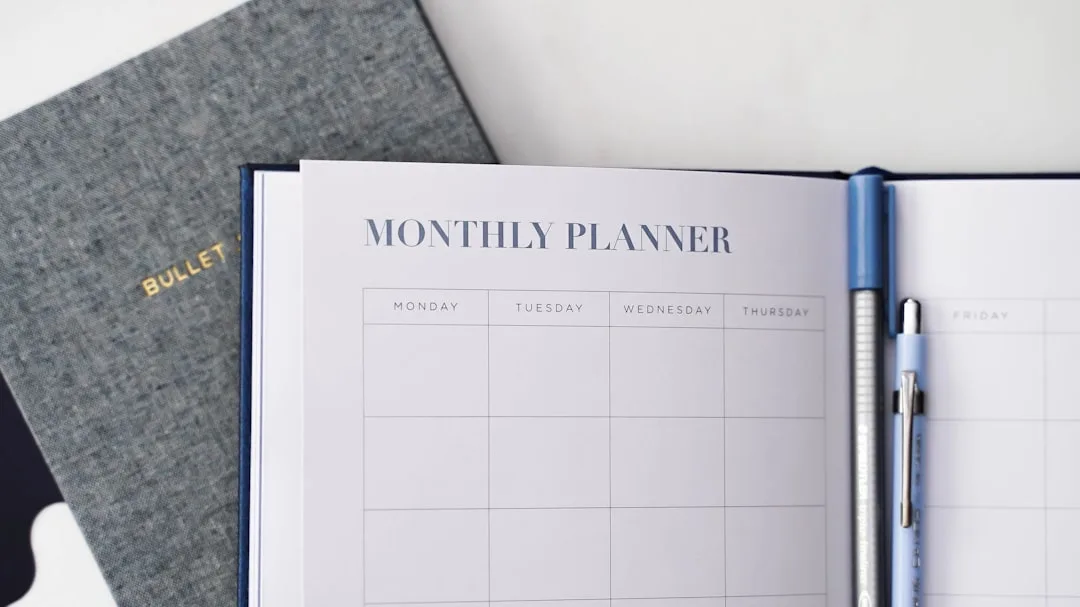 Open monthly planner notebook with weekly grid layout beside two pens, next to a bullet journal on white surface