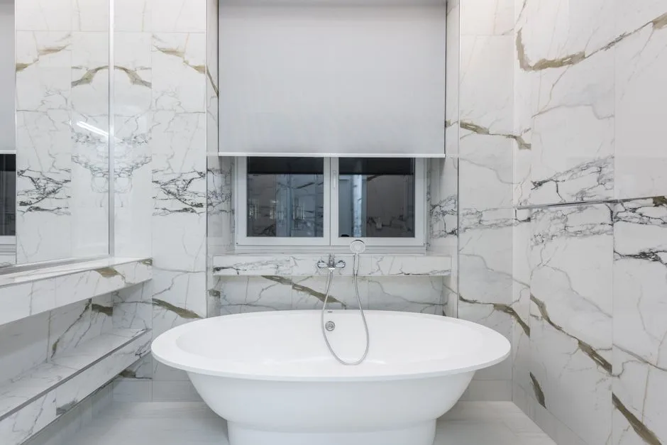 Freestanding oval soaking tub in small marble bathroom with window and roller blind