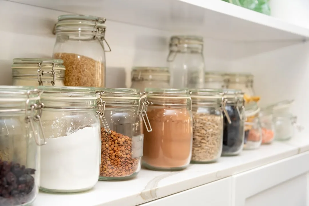 Glass pantry jars organized on white shelves with lentils, flour, cocoa, oats and grains grouped by category