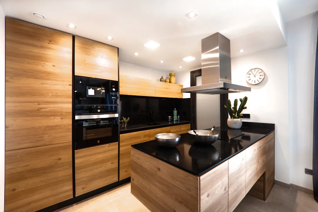 Modern kitchen with warm wood cabinets, black countertops, recessed lighting and stainless steel range hood island