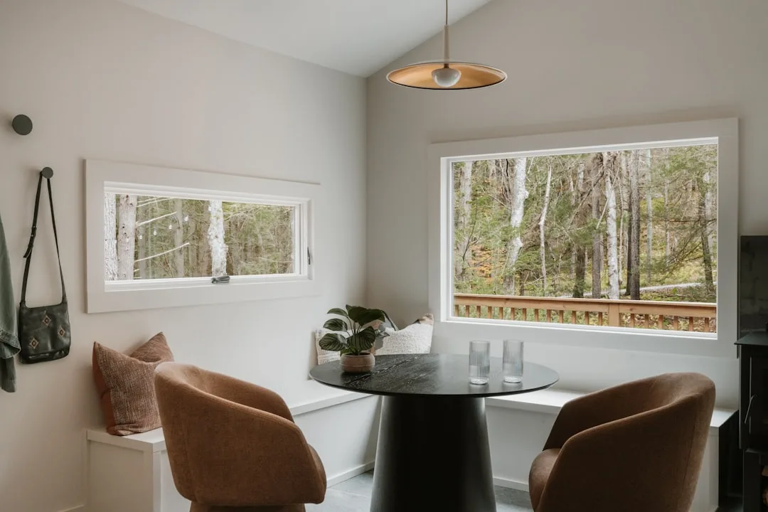 Modern built-in banquette dining nook with black round table, brown chairs, and forest views through large windows