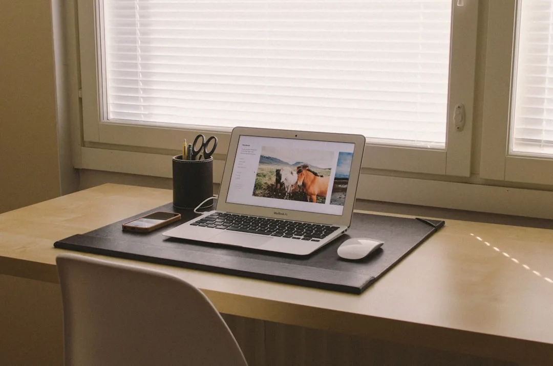 MacBook Air on a black desk mat placed on a shallow wooden desk surface near a window with blinds