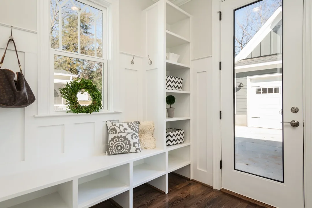 White mudroom entryway with built-in bench, open cubbies, shelving, coat hooks, and glass-panel door in compact space