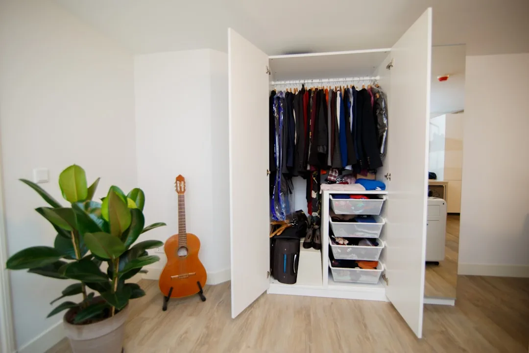 Open white wardrobe closet with hanging clothes and storage drawers in a small bedroom with wood floors