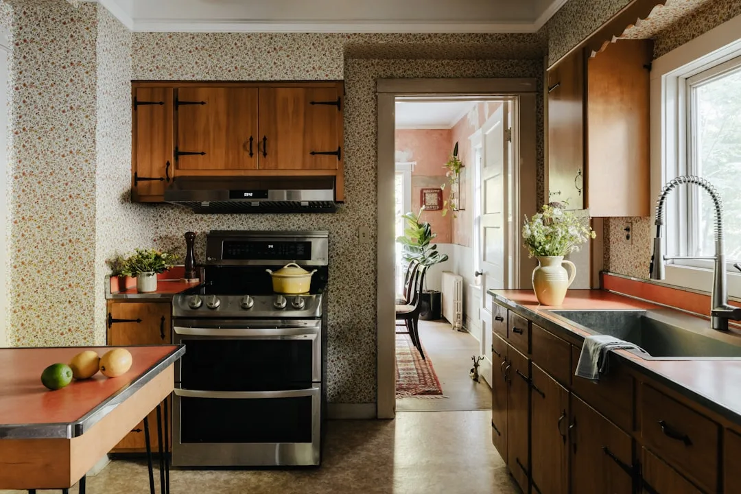 Vintage galley kitchen with floral wallpaper, stainless steel double oven, wood cabinets, and farmhouse sink
