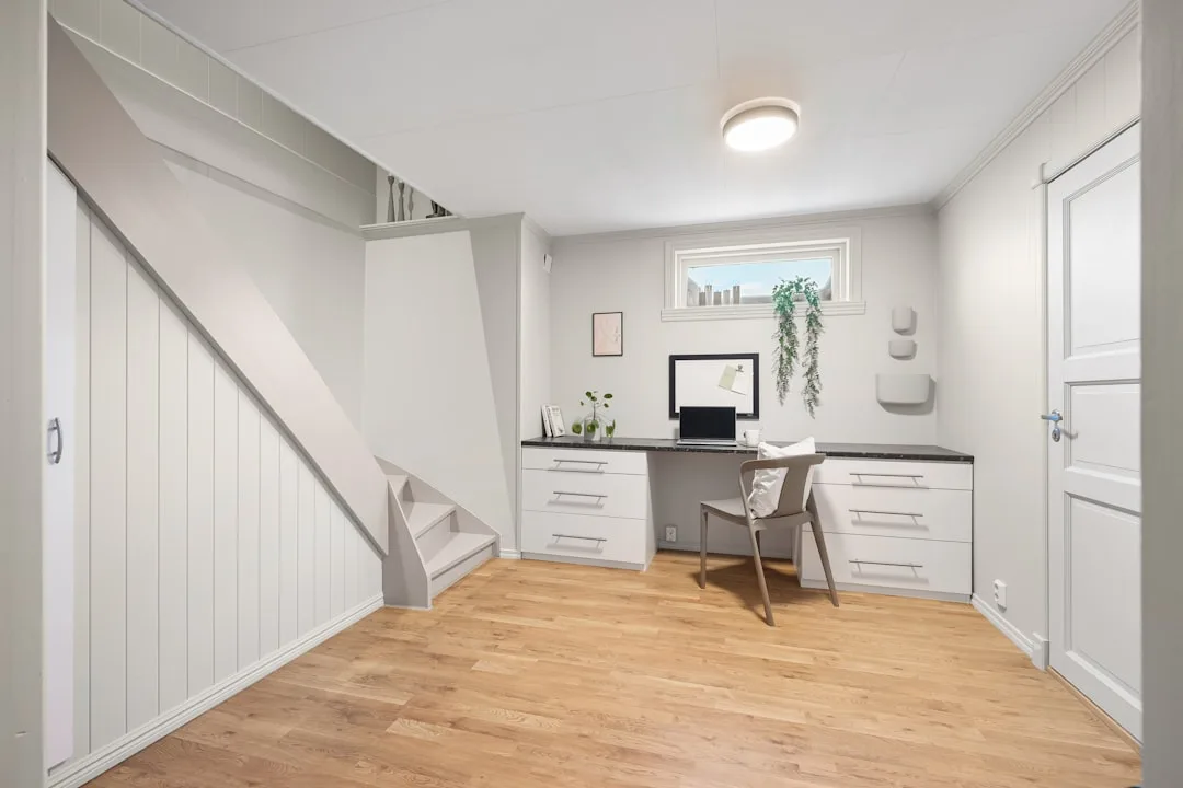 Under-stair home office nook with white built-in drawers, dark countertop desk, wood floors, and small window