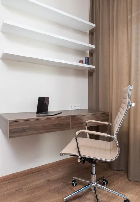 Floating walnut desk with shallow surface mounted on white wall with shelves above and modern office chair