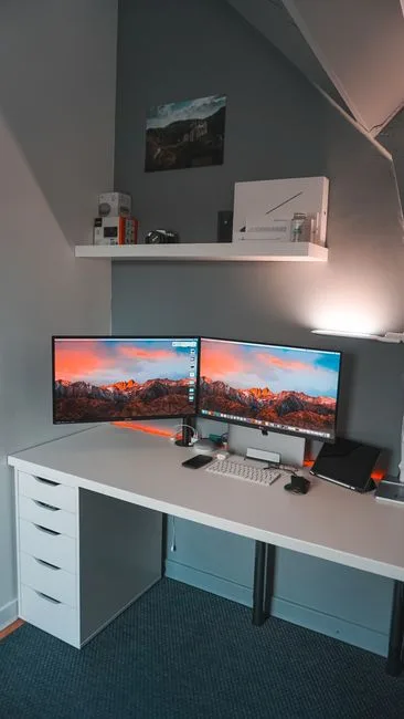 Dual monitor setup on white L-shaped desk with two 24-inch displays showing mountain wallpaper in home office
