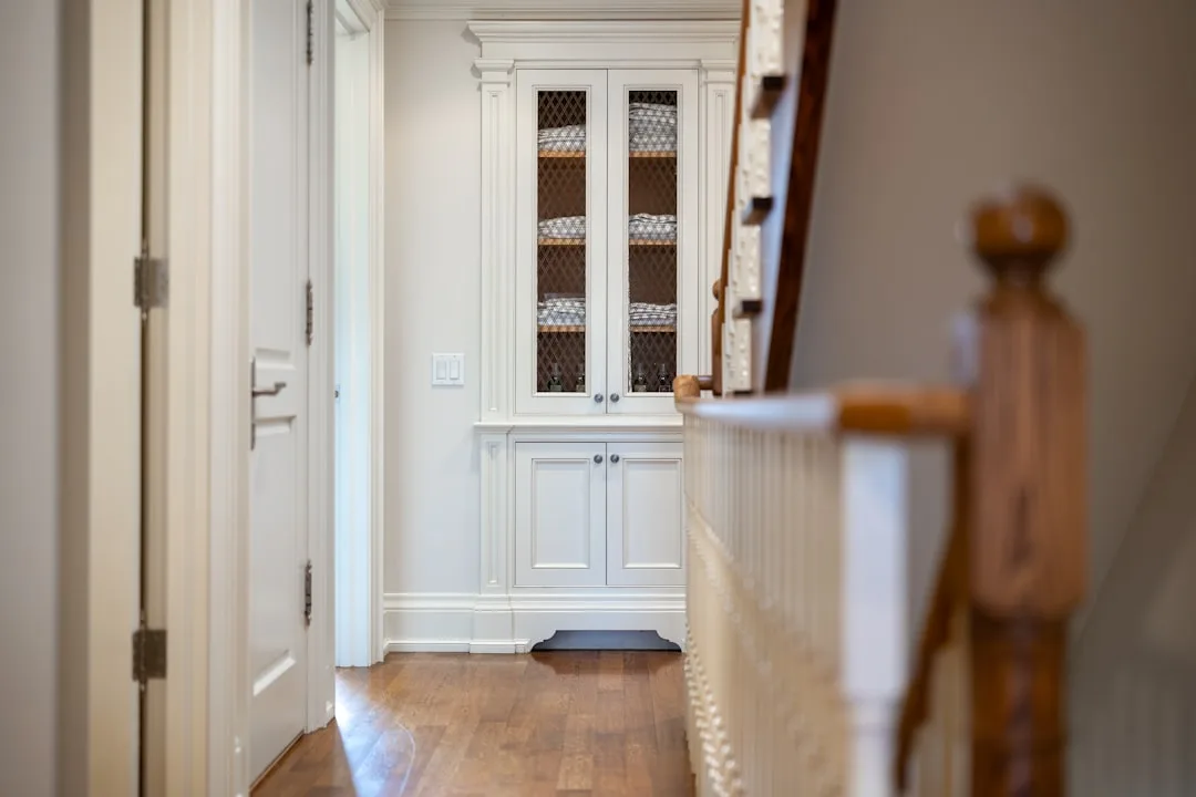 White built-in hallway cabinet with mesh glass doors storing folded linens beside wooden staircase railing