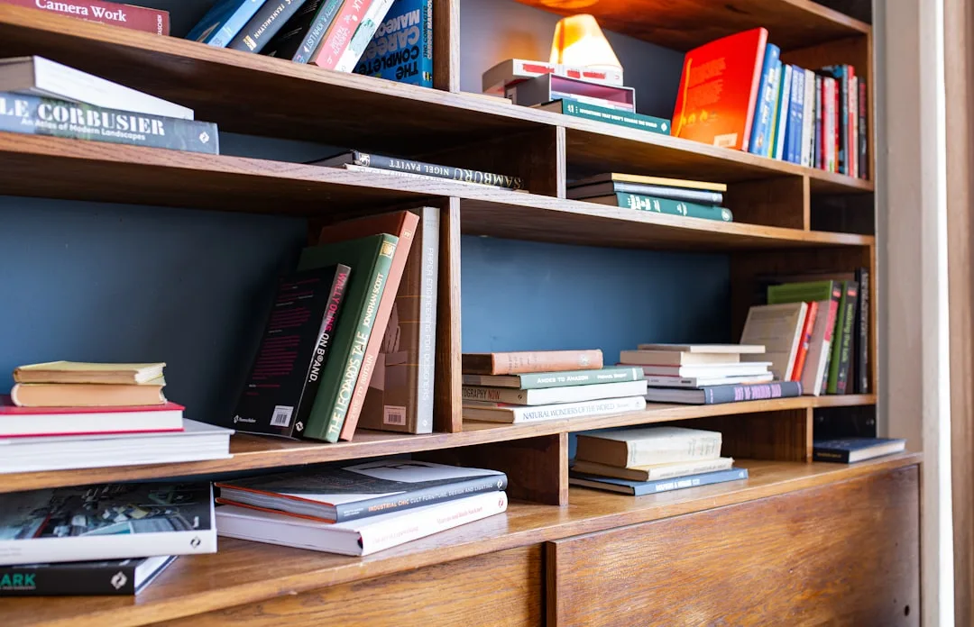 Wooden home office bookshelf with multiple tiers displaying grouped books, decorative objects, and a small lamp