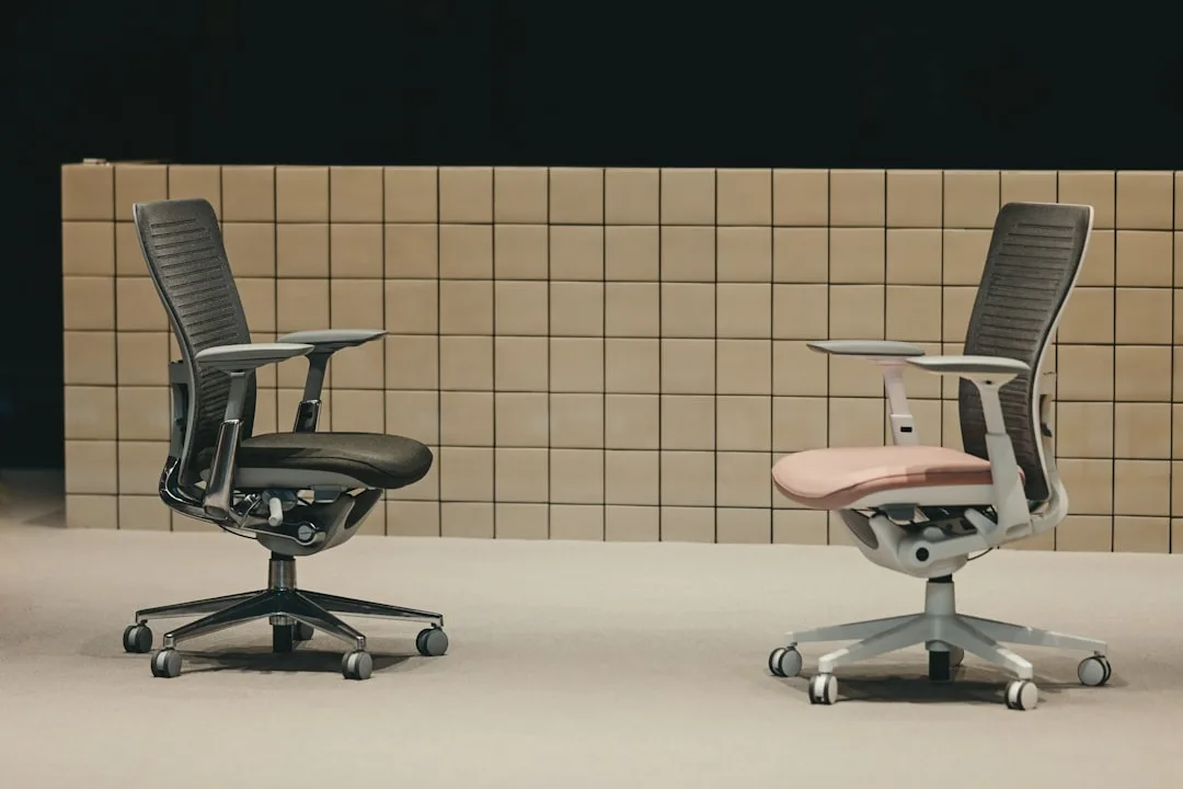 Two ergonomic office chairs side by side — black mesh and white frame with pink seat — showing height-adjustable design