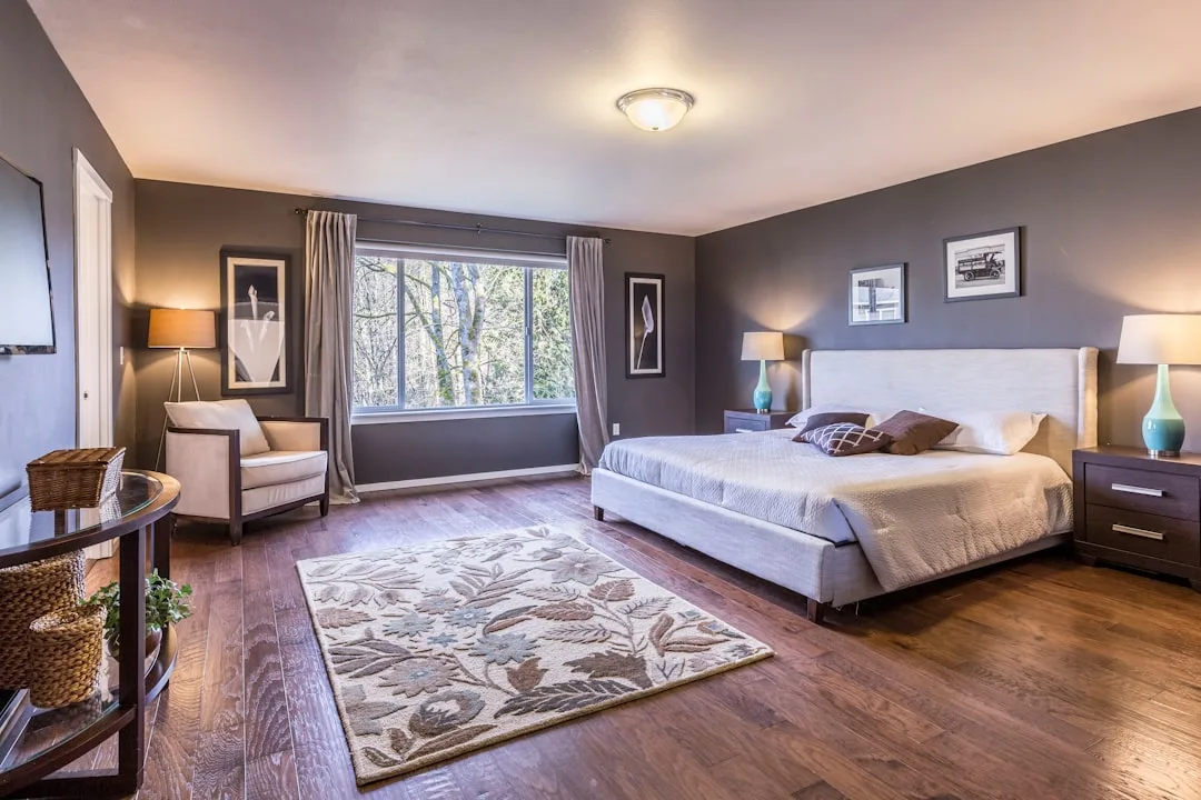 Spacious master bedroom with hardwood floors, gray walls, upholstered bed, layered rugs, and ambient lamps