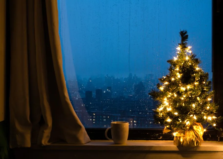Small Christmas tree with warm lights on apartment windowsill with city skyline view at night