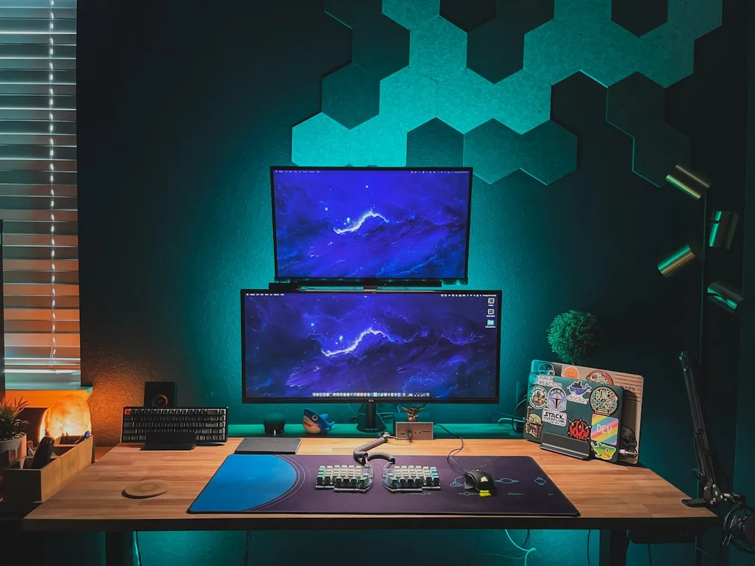 Dual monitor gaming desk setup with hexagon acoustic panels, teal LED lighting, and sticker-covered laptop on wooden des