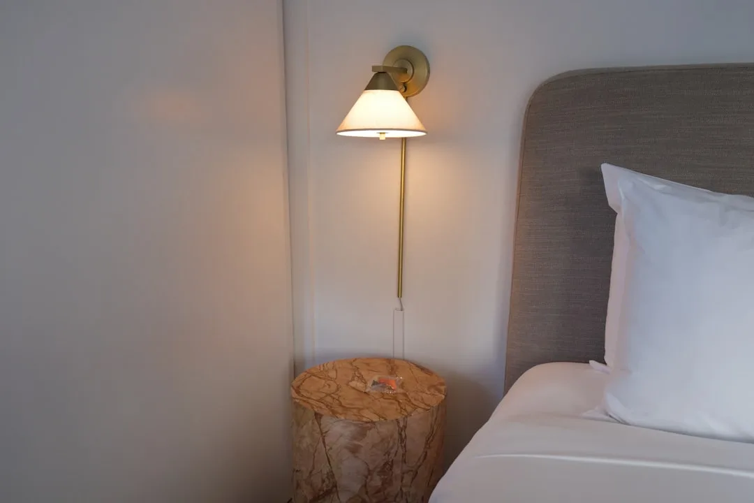 Brass wall sconce with white shade mounted above marble nightstand beside upholstered bed headboard in modern bedroom
