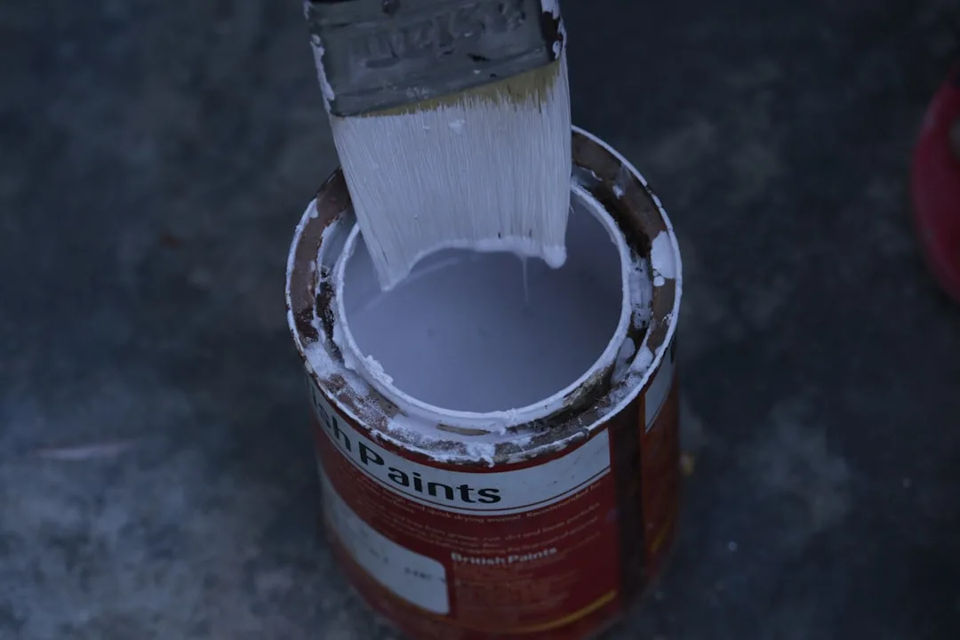 Paintbrush dipping into an open can of white-gray British Paints enamel, showing paint color and undertones
