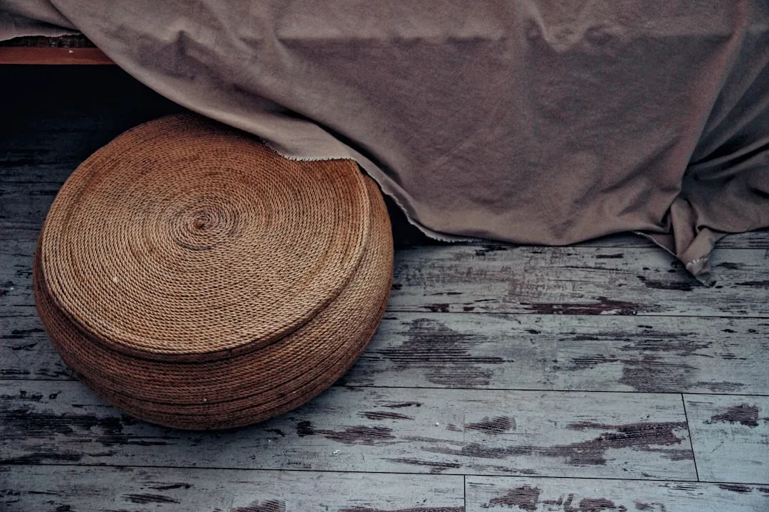 Round woven rattan ottoman on distressed wood floor beside draped fabric, showing drum-like appearance without styling