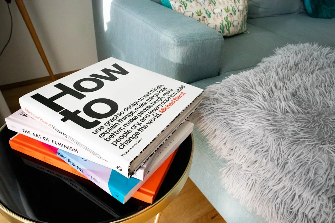 Stack of design and art books on a round side table next to a teal sofa with fluffy throw pillow
