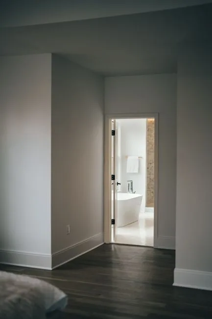 View through bedroom doorway into bright white bathroom with freestanding soaking tub and tile walls